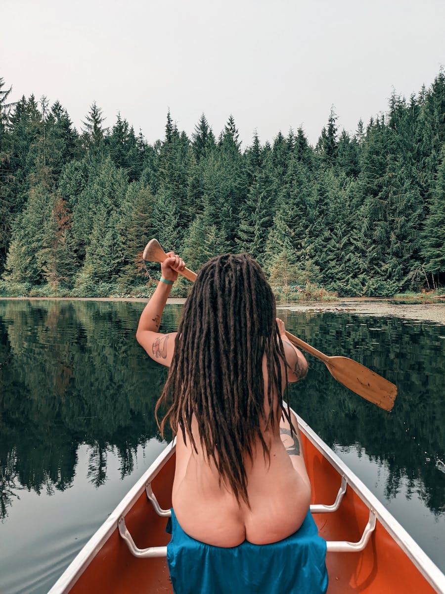 Dreadhead young naked woman is paddling in red color kayak on calm lake in summer